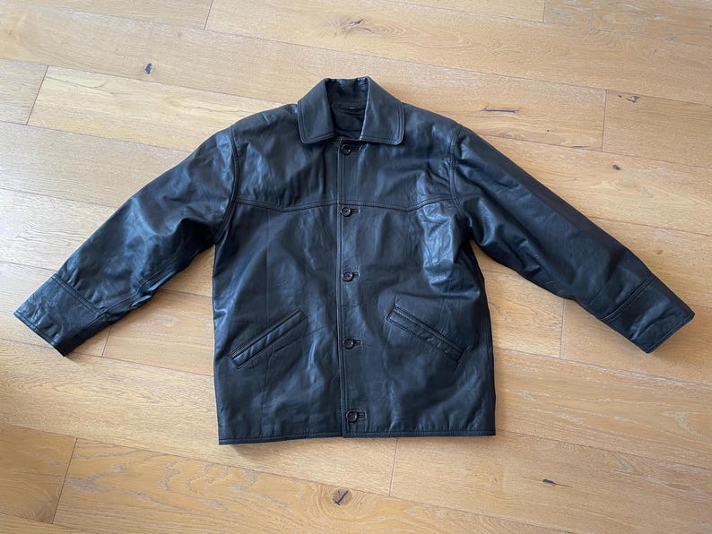 Pu&ograve; includere: Giacca in pelle nera con chiusura a bottoni, due tasche frontali e colletto. La giacca &egrave; stesa su una superficie di legno, mostrando il suo design e la sua consistenza. Le maniche della giacca sono estese.