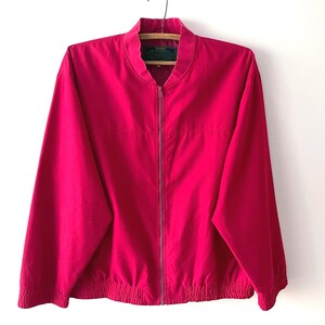 Könnte beinhalten: Eine leuchtend magentafarbene Bomberjacke mit durchgehendem Frontreißverschluss, Stehkragen sowie elastischen Bündchen an Ärmeln und Saum. Die Jacke hat lange Ärmel und besteht aus einem glatten, einfarbigen Stoff mit einem dunklen Innenetikett.
