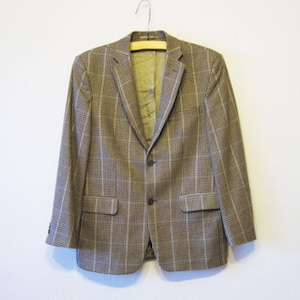 Può includere: Un blazer a quadri marrone e beige con chiusura a tre bottoni e due tasche anteriori. Il blazer è appeso a una gruccia di legno contro un muro bianco. La fodera interna è di colore verde oliva chiaro.