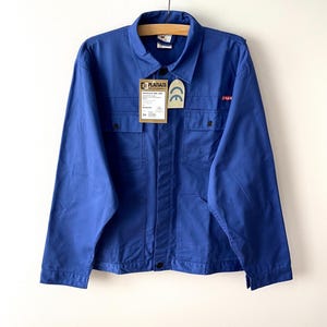 Puede incluir: Una chaqueta de trabajo azul real con cuello clásico, mangas largas y dos bolsillos en el pecho con cierre de botones. La prenda incluye una etiqueta de la marca Planam y una etiqueta de certificación CE, con un pequeño logotipo rojo.