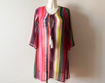 Può includere: Un'abito a tunica a righe colorato con collo annodato e dettagli con nappe. L'abito presenta righe verticali nelle tonalità del rosa, rosso, verde e bianco. Le maniche sono a tre quarti.