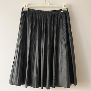 May include: A dark gray pleated skirt hanging on a white hanger. The skirt has a button detail at the waistband. The fabric appears to have a subtle sheen. The skirt is a midi length.