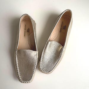 Peut inclure: Une paire de mocassins dorés métallisés avec une finition texturée et des coutures visibles le long de l'empeigne. L'intérieur des chaussures présente une doublure claire avec le nom de marque Peter Hahn imprimé en noir sur le talon.