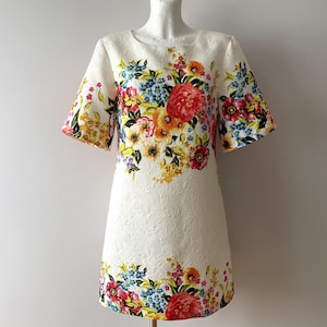 Peut inclure: Robe blanche à imprimé floral avec manches courtes. La robe présente une encolure ronde et un ourlet au genou. Le motif floral comprend des fleurs rouges, jaunes, bleues et oranges. Le tissu a un motif texturé subtil.