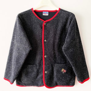 May include: A dark gray cardigan with red trim and buttons. The cardigan has two front pockets and a small floral embroidery on one side. The collar and edges are trimmed in red. The garment is hanging on a white hanger.