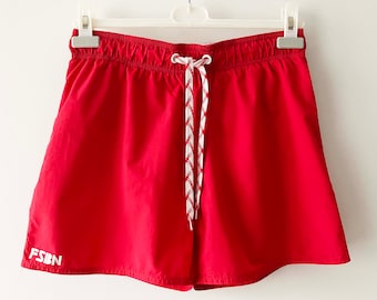 Könnte beinhalten: Leuchtend rote Sporthose mit weißem Kordelzug und dem weißen Schriftzug "FSBN" am linken Bein. Die Shorts hängen an einem weißen Kleiderbügel. Die Shorts sind aus leichtem Material gefertigt und eignen sich für Sportaktivitäten.