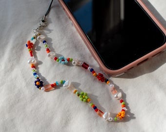 Op de afbeelding: Een kleurrijke, met kralen versierde telefoonbedel met parels, bloemvormige kralen en heldere rechthoekige kralen. De bedel is bevestigd aan een roze telefoonhoesje. De kralen zijn in tinten rood, oranje, geel, groen, blauw en wit.