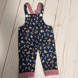May include: Navy blue toddler overalls with a colorful mushroom print. The straps and cuffs are lined with red and white gingham fabric. Light blue buttons secure the straps.