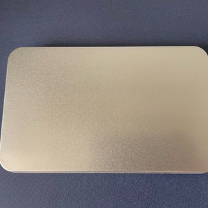 May include: A silver metal rectangular tin box with a smooth, slightly textured surface.