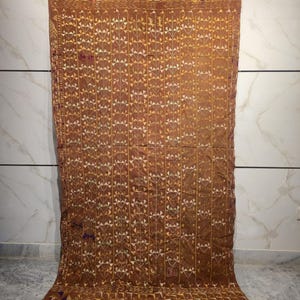May include: A brown textile with a geometric pattern of squares and triangles. The textile is hanging from a metal rod with clips. The pattern is in shades of yellow and white, with some purple accents.