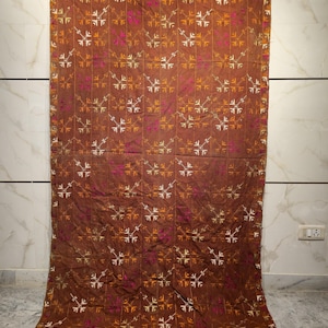 May include: A brown textile featuring a repeating pattern of white, pink, and orange geometric shapes. The fabric is displayed vertically, showcasing its full length. The textile is likely a blanket or wall hanging.