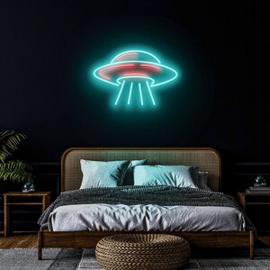 Puede incluir: Un letrero de ne&oacute;n azul verdoso con forma de platillo volante con un interior rojo. El letrero est&aacute; montado en una pared azul oscuro sobre una cama con un marco de madera y una manta gris.