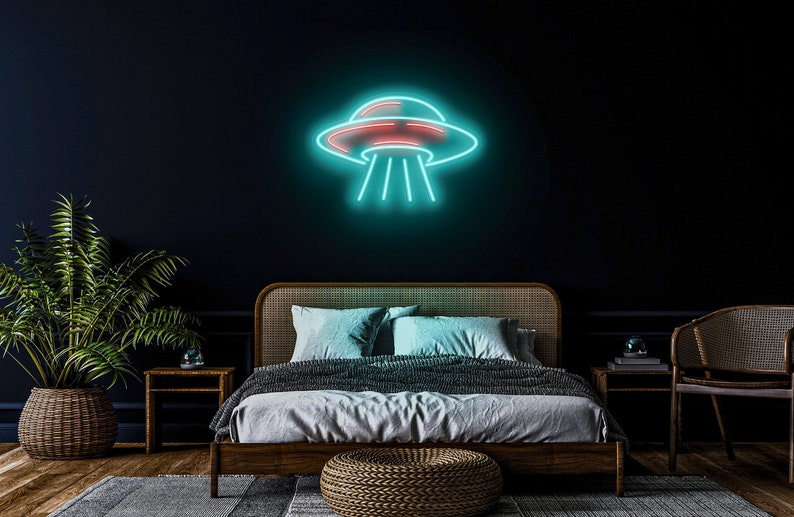 Puede incluir: Un letrero de ne&oacute;n azul verdoso con forma de platillo volante con un interior rojo. El letrero est&aacute; montado en una pared azul oscuro sobre una cama con un marco de madera y una manta gris.