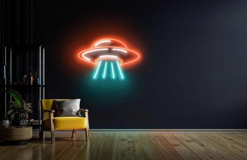 Puede incluir: Un letrero de ne&oacute;n con forma de platillo volante con luces naranja y azules sobre una pared oscura. El letrero est&aacute; colgado sobre un sill&oacute;n amarillo en una sala de estar.