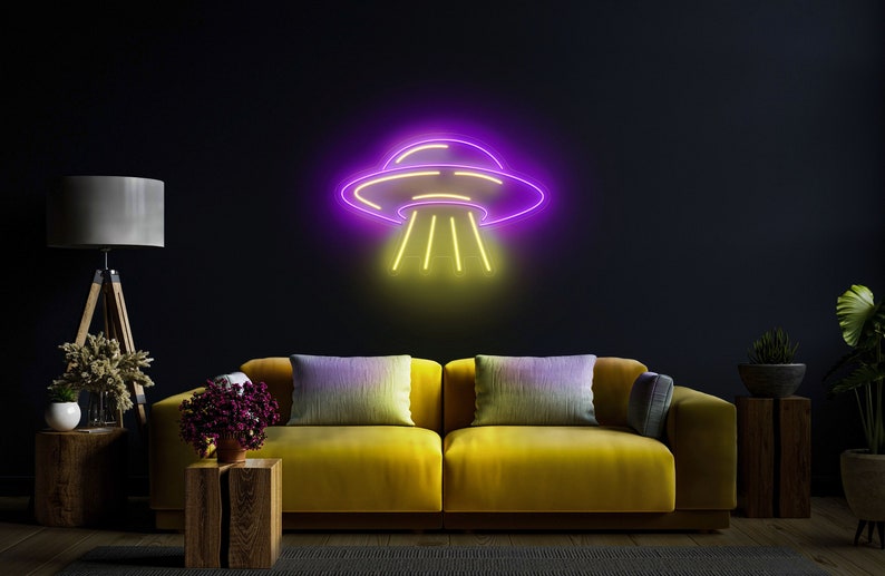 Puede incluir: Un letrero de ne&oacute;n con forma de platillo volante con un contorno morado y luces amarillas. El letrero est&aacute; colgado en una pared oscura sobre un sof&aacute; amarillo con almohadas.