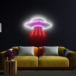 Puede incluir: Un letrero de ne&oacute;n con forma de platillo volante con luces rosas y blancas. El letrero est&aacute; montado en una pared oscura sobre un sof&aacute; amarillo con almohadas grises.