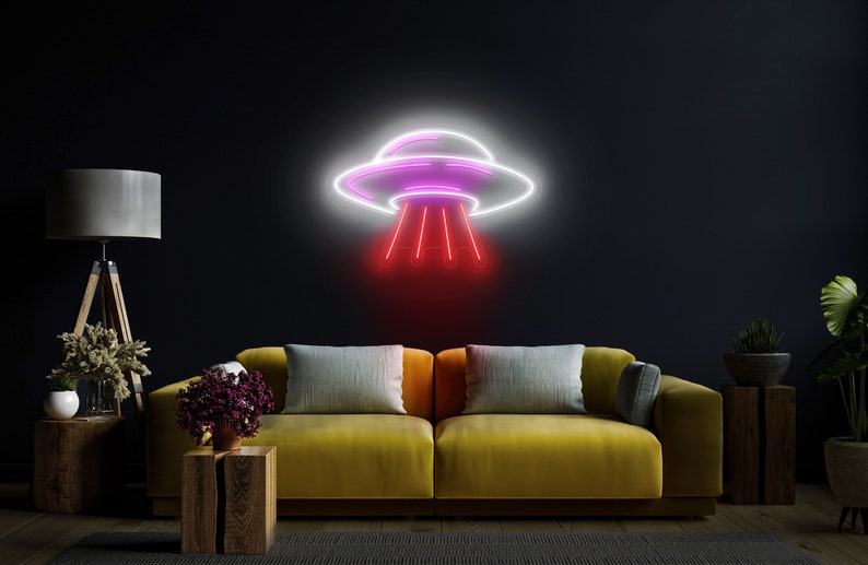 Puede incluir: Un letrero de ne&oacute;n con forma de platillo volante con luces rosas y blancas. El letrero est&aacute; montado en una pared oscura sobre un sof&aacute; amarillo con almohadas grises.