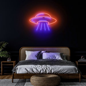 Puede incluir: Un letrero de ne&oacute;n con forma de platillo volante con un brillo morado y naranja. El letrero est&aacute; colgado en una pared oscura sobre una cama con un marco de madera y una manta gris.