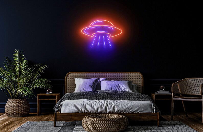 Puede incluir: Un letrero de ne&oacute;n con forma de platillo volante con un brillo morado y naranja. El letrero est&aacute; colgado en una pared oscura sobre una cama con un marco de madera y una manta gris.