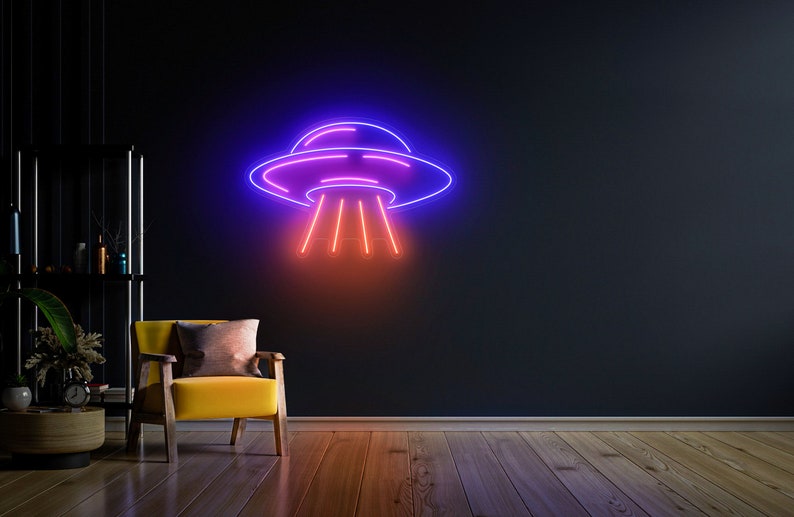 Puede incluir: Un letrero de ne&oacute;n con forma de platillo volante con un contorno morado y azul y rayos de luz rojos. El letrero est&aacute; montado en una pared oscura en una habitaci&oacute;n con un sill&oacute;n amarillo.