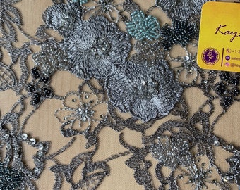 Puede incluir: Tela de encaje floral con cuentas plateadas y azules. La tela presenta una base de malla delicada con flores bordadas y cuentas.