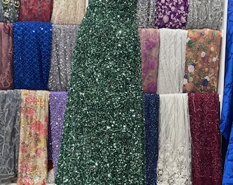 May include: A green sequined fabric with a one-shoulder design. The fabric is displayed on a mannequin and surrounded by other fabrics.