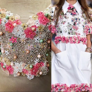 以下が含まれることがあります： 花柄の白いドレス。ドレスはピンク、白、緑の花と真珠で覆われています。ドレスは襟と半袖です。