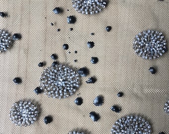 Puede incluir: Primer plano de una tela beige con adornos negros y plateados. La tela tiene una textura de malla y está cubierta de pequeñas cuentas redondas plateadas que están dispuestas en un patrón circular. También hay puntos negros dispersos por toda la tela.