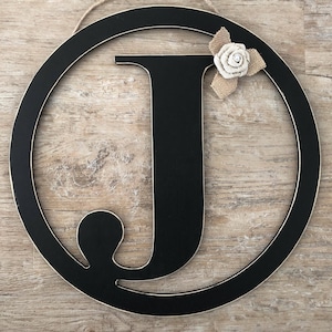 May include: Black wooden letter J in a circular frame with a burlap and white flower accent.