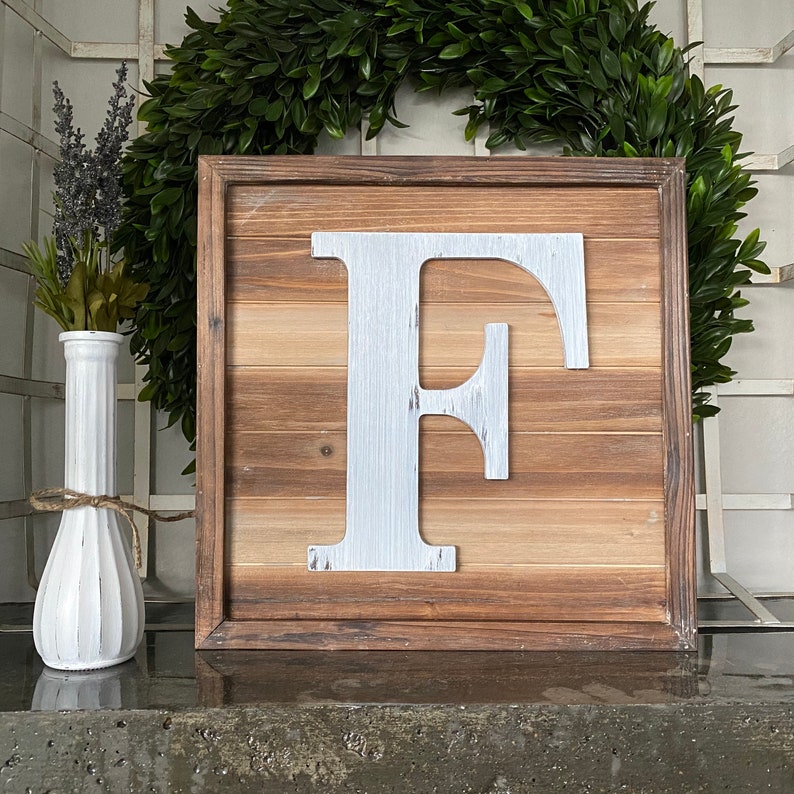 May include: A wooden sign with a white painted letter F. The sign has a rustic wood grain and is framed in a dark brown wood frame.