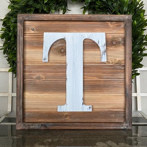 May include: A wooden framed sign with a white painted letter "T" in the center. The wood has a rustic, distressed look.