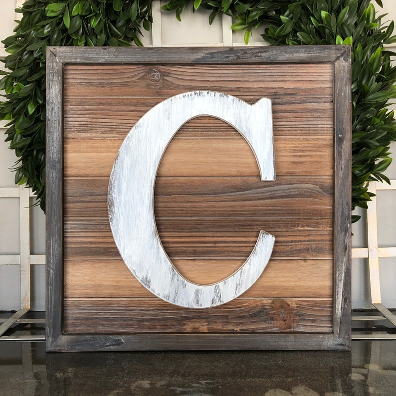 May include: A wooden sign with a distressed brown finish and a white painted letter "C" in the center. The sign has a dark brown frame.