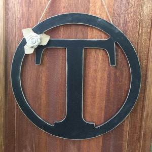 May include: Black wooden letter T in a circular frame with a small white flower accent. The letter is hanging from a string.