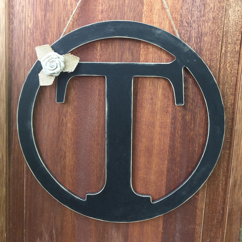 May include: Black wooden letter T in a circular frame with a small white flower accent. The letter is hanging from a string.