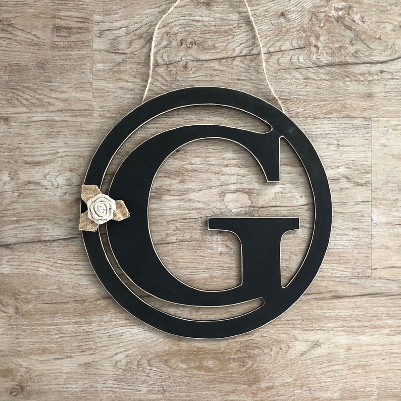 May include: Black wooden letter G with a burlap and white fabric flower attached. The letter is hanging from a string.