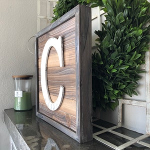 May include: A wooden sign with a white letter "C" on it. The sign has a dark wood frame and is made of reclaimed wood.