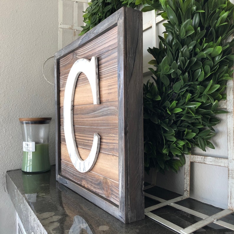 May include: A wooden sign with a white letter "C" on it. The sign has a dark wood frame and is made of reclaimed wood.