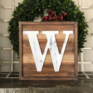 May include: A white wooden letter W in a rustic brown wood frame. The letter is distressed and has a whitewashed finish. The frame is made of reclaimed wood and has a natural finish.