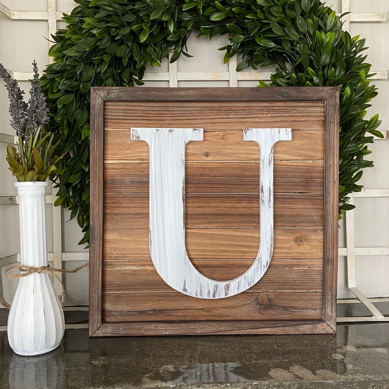 May include: A wooden sign with a white painted letter U. The sign is brown with a rustic wood grain texture. The letter U is centered on the sign and is painted in a distressed white paint.