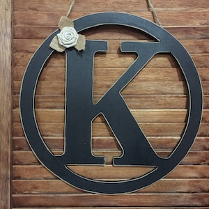 May include: Black wooden letter K in a circular frame with a burlap and white flower embellishment.