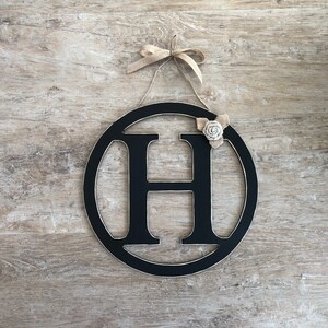 May include: Black wooden circle with the letter H cut out. The circle is decorated with a burlap bow and a small white fabric flower.
