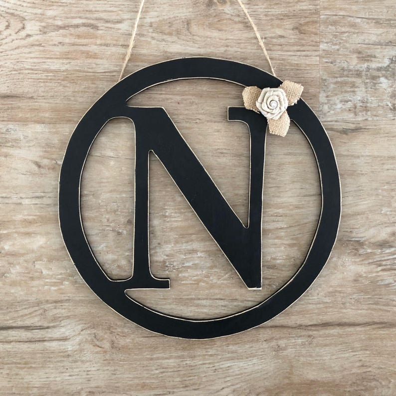May include: Black wooden circle with a black letter N in the center. The circle has a burlap bow with a white flower on top.