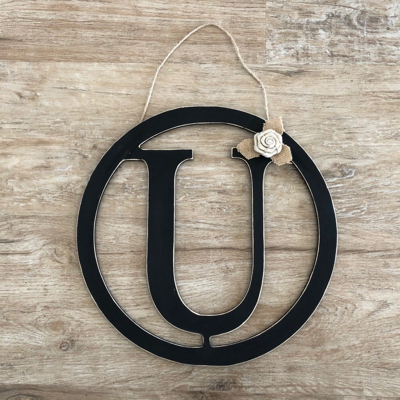 May include: Black wooden letter U with a burlap and white fabric flower attached. The letter is in a circular frame and has a string for hanging.