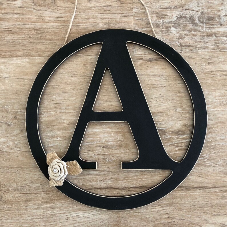 May include: Black wooden circle with the letter A in a white outline. A small burlap flower with a white center is attached to the bottom of the circle.