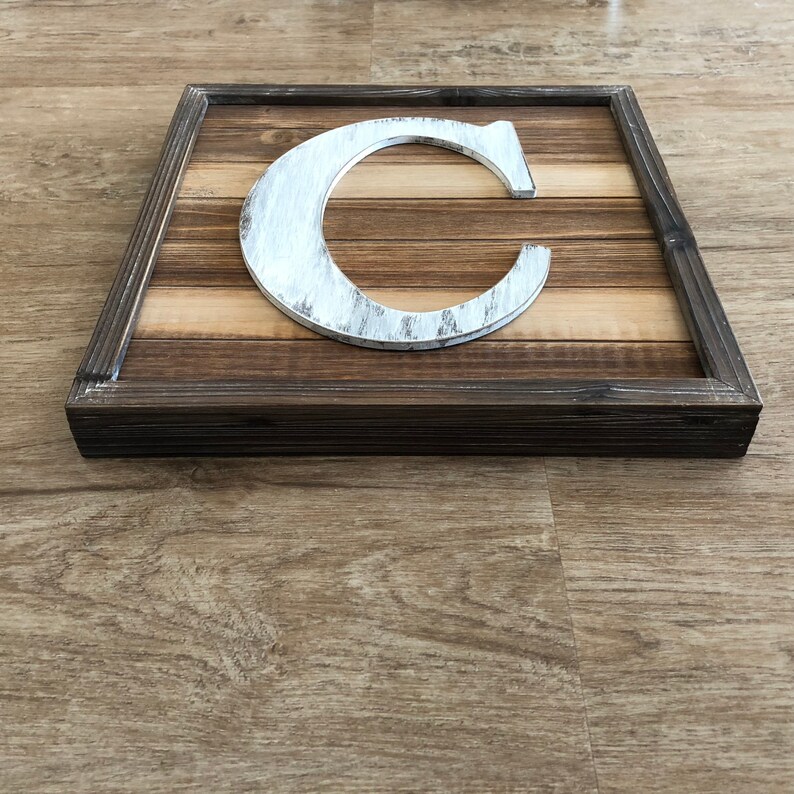May include: A white wooden letter C with a distressed finish is displayed in a rustic wooden frame. The frame has a dark brown stain and is made of multiple wood planks.