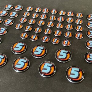 Puede incluir: Una colecci&oacute;n de fichas de juego con los n&uacute;meros 1, 3 y 5. Las fichas son redondas y tienen un acabado de metal plateado. Los n&uacute;meros est&aacute;n impresos en blanco sobre un fondo azul con llamas naranjas.