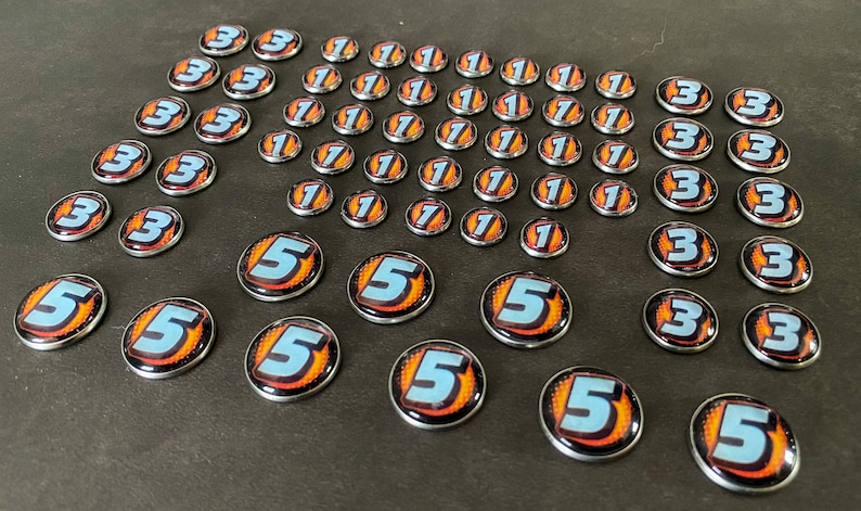 Puede incluir: Una colecci&oacute;n de fichas de juego con los n&uacute;meros 1, 3 y 5. Las fichas son redondas y tienen un acabado de metal plateado. Los n&uacute;meros est&aacute;n impresos en blanco sobre un fondo azul con llamas naranjas.