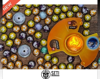 Könnte beinhalten: Ein Brettspiel mit einem gelben, orangefarbenen und weißen Spielbrett mit den Worten "Merkur", "Venus" und "11" darauf. Das Spielbrett ist von kleinen, bunten Spielsteinen mit Symbolen und Zahlen umgeben. Das Spiel heißt "SETI".