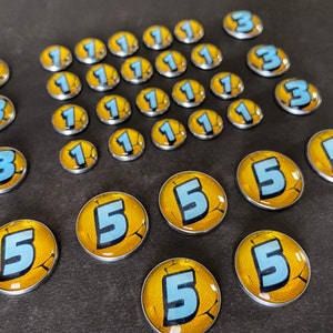 Puede incluir: Una colecci&oacute;n de fichas redondas de color plateado con dise&ntilde;os amarillos y azules. Las fichas presentan los n&uacute;meros 1, 3 y 5.