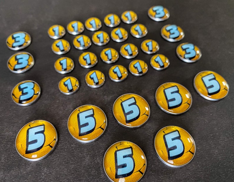 Puede incluir: Una colecci&oacute;n de fichas redondas de color plateado con dise&ntilde;os amarillos y azules. Las fichas presentan los n&uacute;meros 1, 3 y 5.
